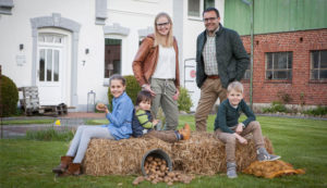 Bild: Familie vor Hof, Kinder sitzen auf Strohballen umgeben von Kartoffeln im Sack und Eimer